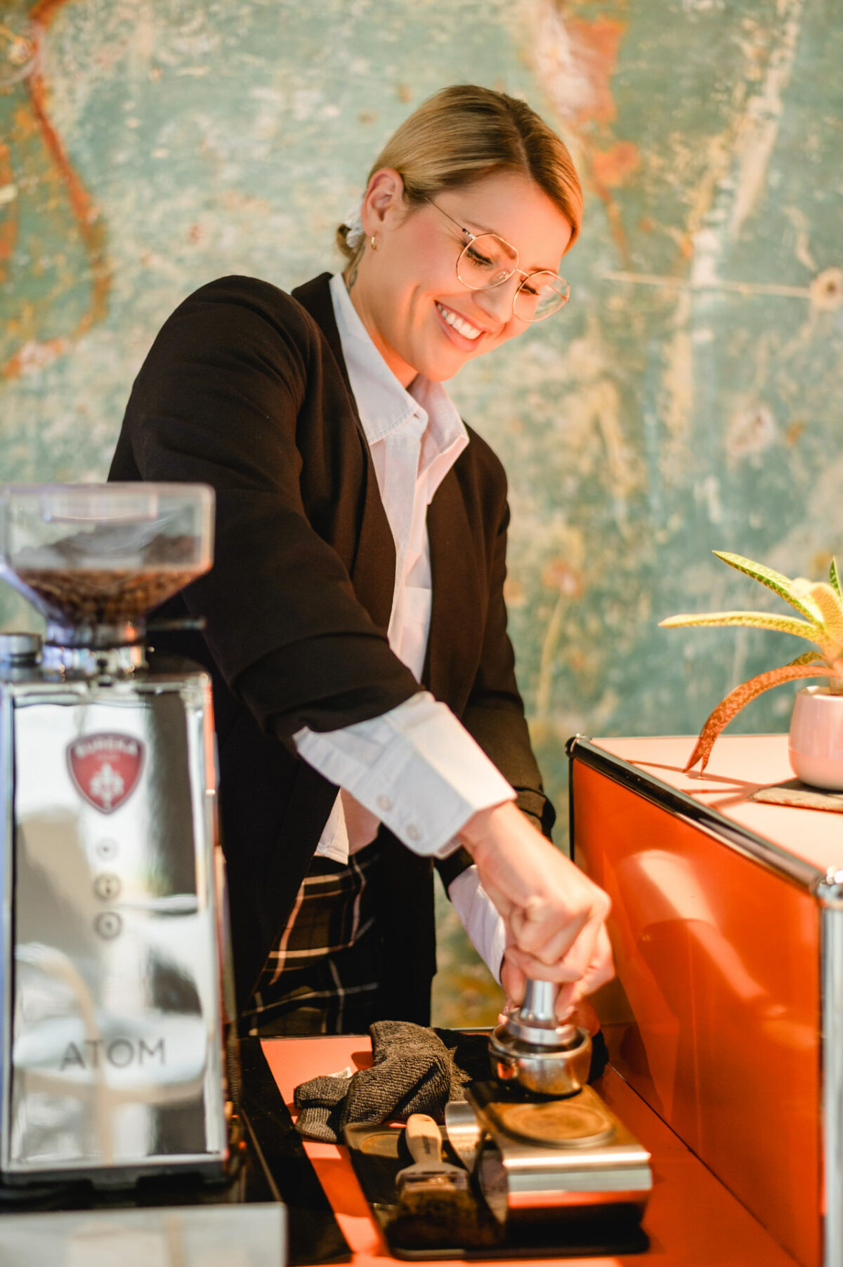 Christian Lorenz Fotograf Eine lächelnde Frau mit Brille und Blazer presst gemahlenen Kaffee mit einem Tamper neben einer Kaffeemühle und Werkzeugen auf einer orangefarbenen Theke, mit einer grün strukturierten Wand im Hintergrund. Christian Lorenz Fotograf Eine lächelnde Frau mit Brille und Blazer presst gemahlenen Kaffee mit einem Tamper neben einer Kaffeemühle und Werkzeugen auf einer orangefarbenen Theke, mit einer grün strukturierten Wand im Hintergrund.