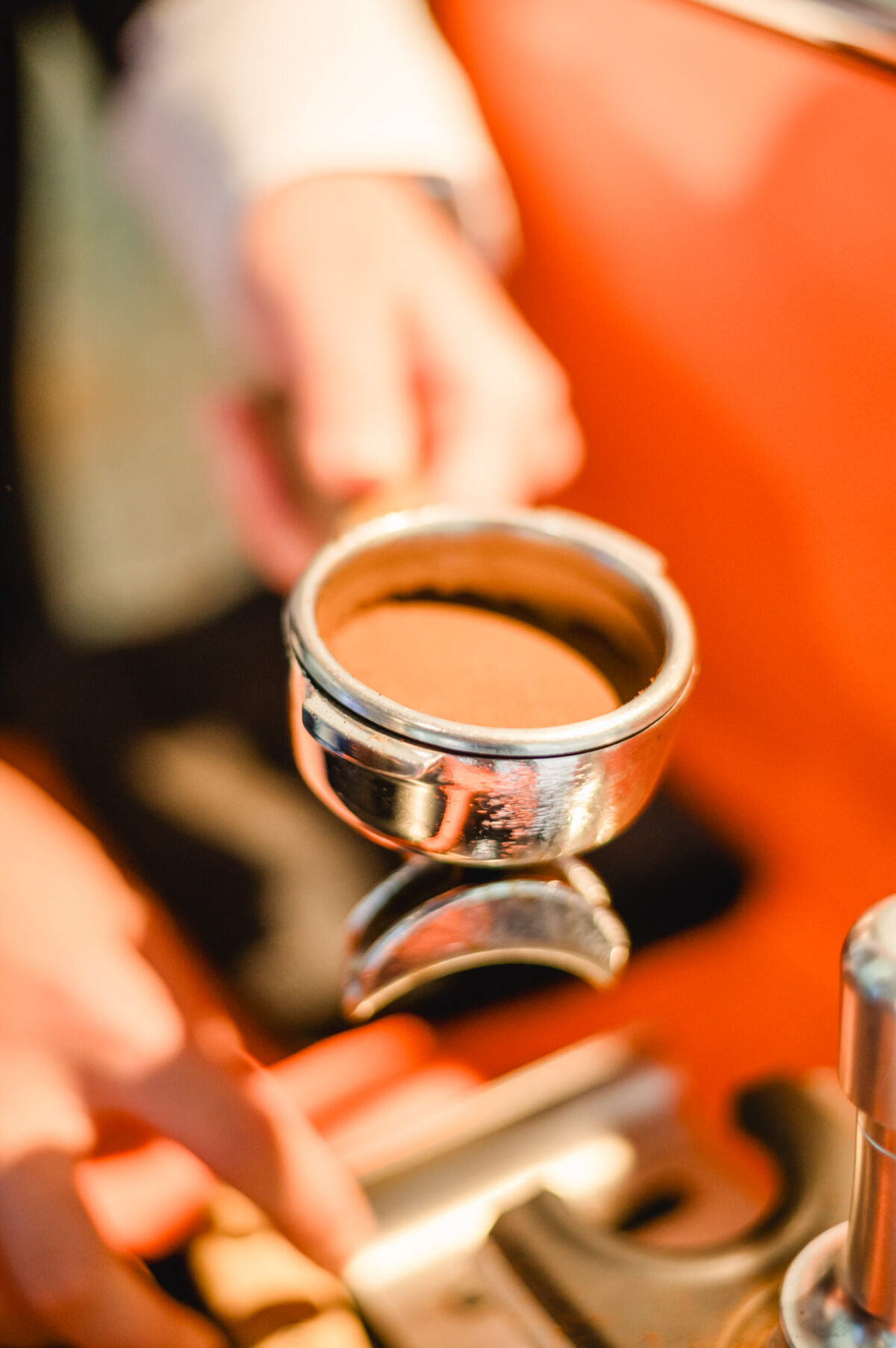 Christian Lorenz Fotograf Eine Person hält einen Tamper und einen mit frisch gemahlenem Kaffee gefüllten Siebträger in der Hand und bereitet sich auf die Zubereitung von Espresso vor, während im Hintergrund eine Espressomaschine zu sehen ist. Christian Lorenz Fotograf Eine Person hält einen Tamper und einen mit frisch gemahlenem Kaffee gefüllten Siebträger in der Hand und bereitet sich auf die Zubereitung von Espresso vor, während im Hintergrund eine Espressomaschine zu sehen ist.