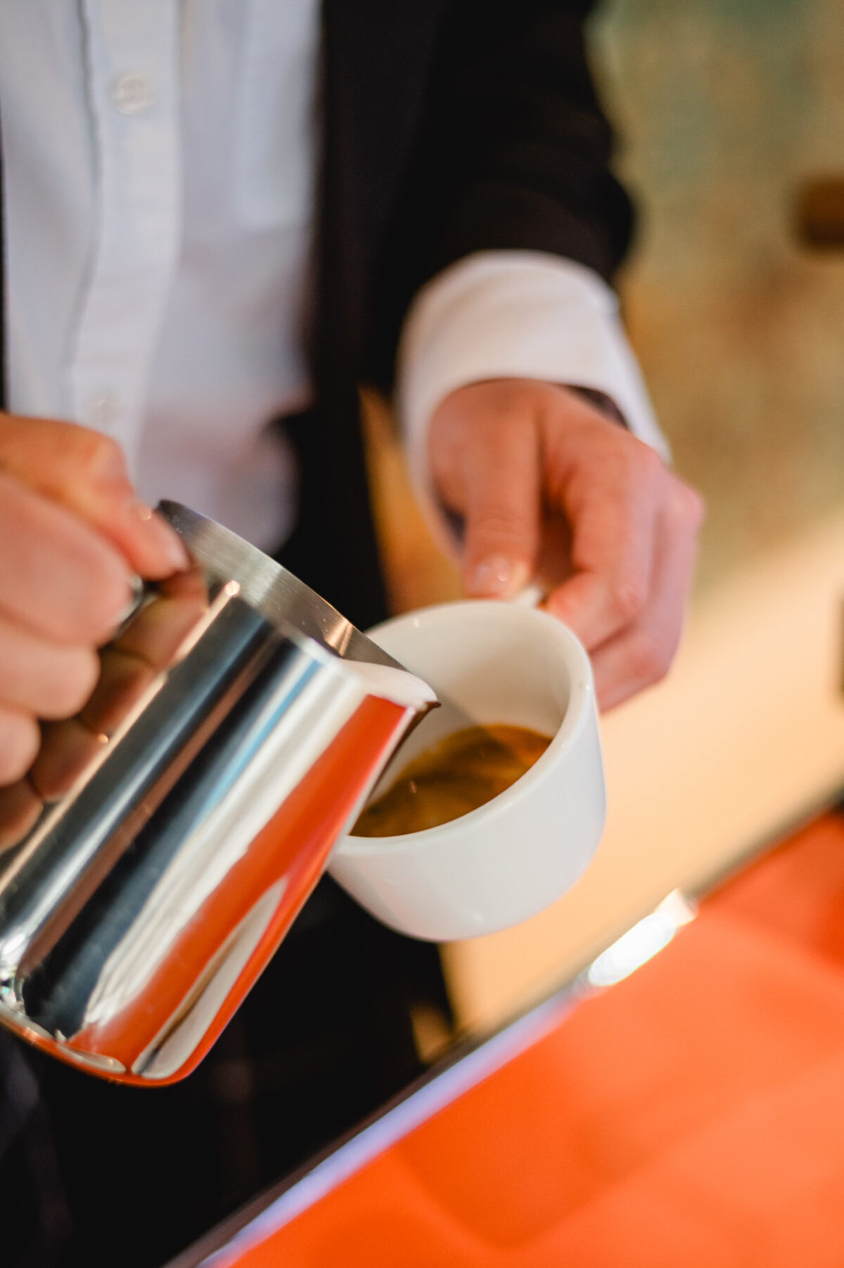 Christian Lorenz Fotograf Eine Person im Anzug gießt gedämpfte Milch aus einer Metallkanne in eine weiße Espressotasse und bereitet ein Kaffeegetränk zu, möglicherweise einen Milchkaffee oder Cappuccino. Christian Lorenz Fotograf Eine Person im Anzug gießt gedämpfte Milch aus einer Metallkanne in eine weiße Espressotasse und bereitet ein Kaffeegetränk zu, möglicherweise einen Milchkaffee oder Cappuccino.