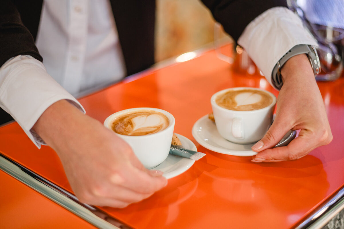 Christian Lorenz Fotograf Eine Person im Anzug serviert zwei Cappuccinos mit Latte Art auf einer leuchtend orangefarbenen Theke und stellt jede Tasse und Untertasse sorgfältig ab. Christian Lorenz Fotograf Eine Person im Anzug serviert zwei Cappuccinos mit Latte Art auf einer leuchtend orangefarbenen Theke und stellt jede Tasse und Untertasse sorgfältig ab.