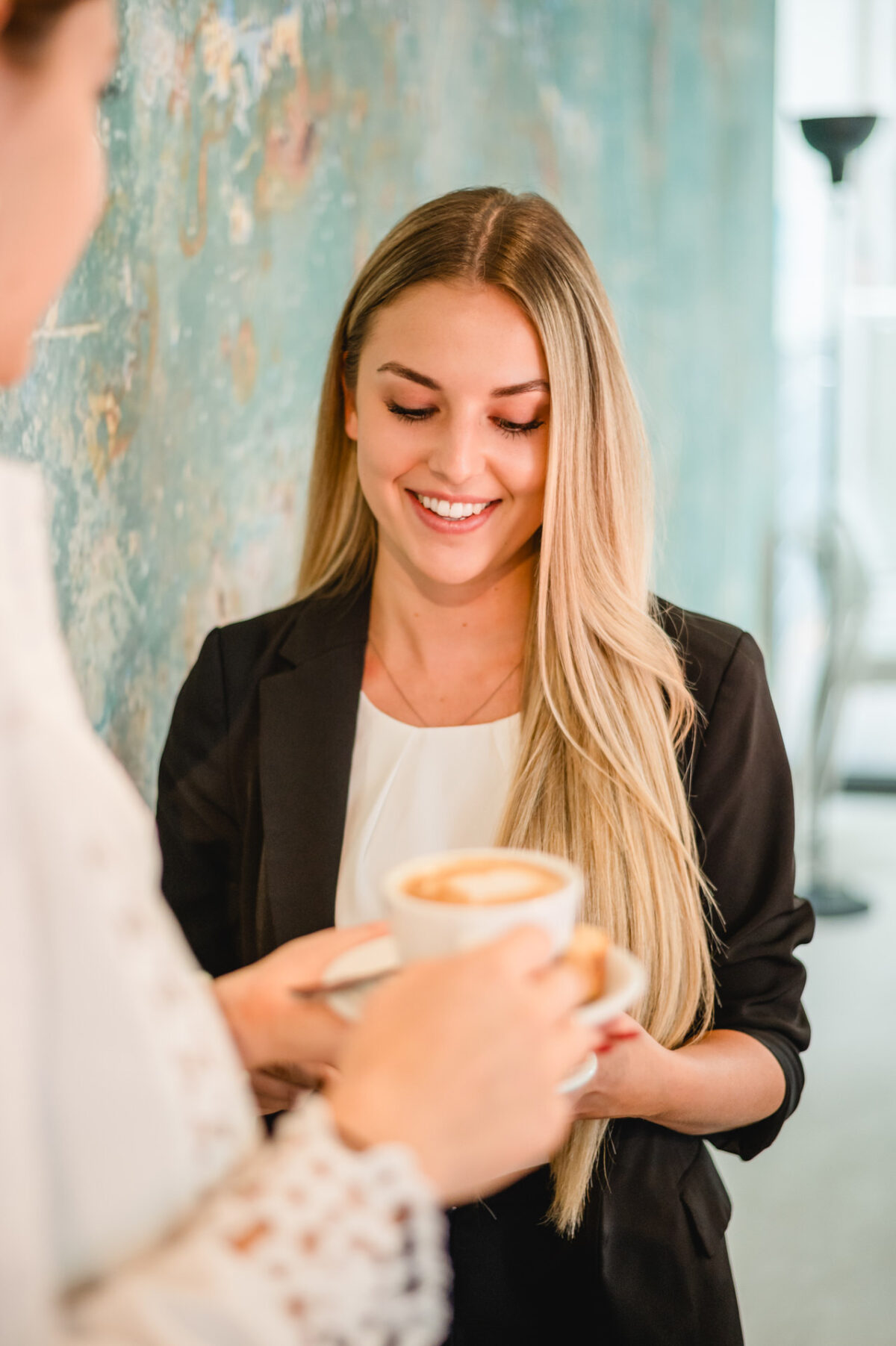 Christian Lorenz Fotograf Eine Frau mit langen blonden Haaren, die einen schwarzen Blazer trägt, lächelt, während sie eine Tasse Kaffee von einer anderen Person in einem schwach beleuchteten Raum mit einer strukturierten tealfarbenen Wand entgegennimmt. Christian Lorenz Fotograf Eine Frau mit langen blonden Haaren, die einen schwarzen Blazer trägt, lächelt, während sie eine Tasse Kaffee von einer anderen Person in einem schwach beleuchteten Raum mit einer strukturierten tealfarbenen Wand entgegennimmt.