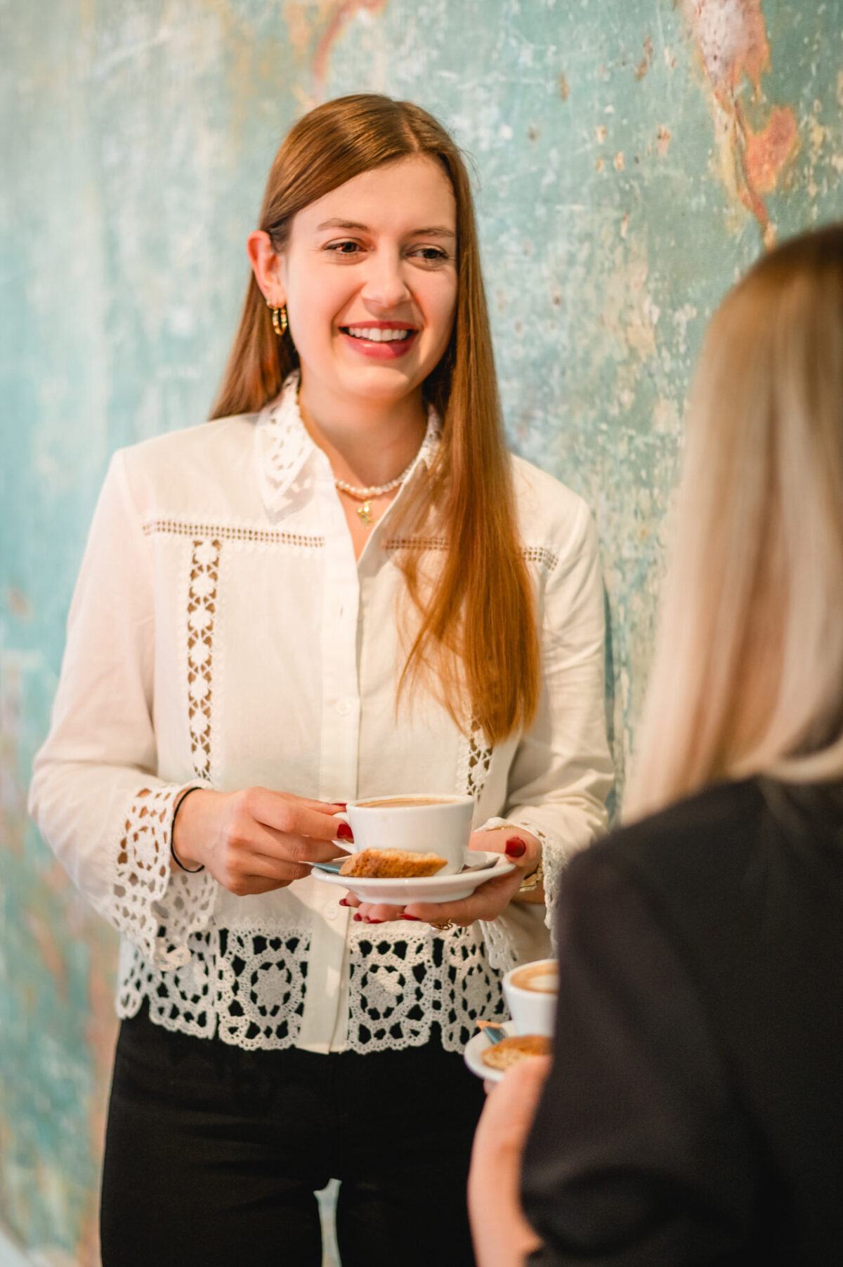 Christian Lorenz Fotograf Eine Frau mit langen braunen Haaren und weißer Bluse lächelt, während sie eine Tasse mit Untertasse hält. Sie steht einer anderen Person mit blondem Haar gegenüber, die sich vor einer grünlichen, strukturierten Wand unterhält. Christian Lorenz Fotograf Eine Frau mit langen braunen Haaren und weißer Bluse lächelt, während sie eine Tasse mit Untertasse hält. Sie steht einer anderen Person mit blondem Haar gegenüber, die sich vor einer grünlichen, strukturierten Wand unterhält.