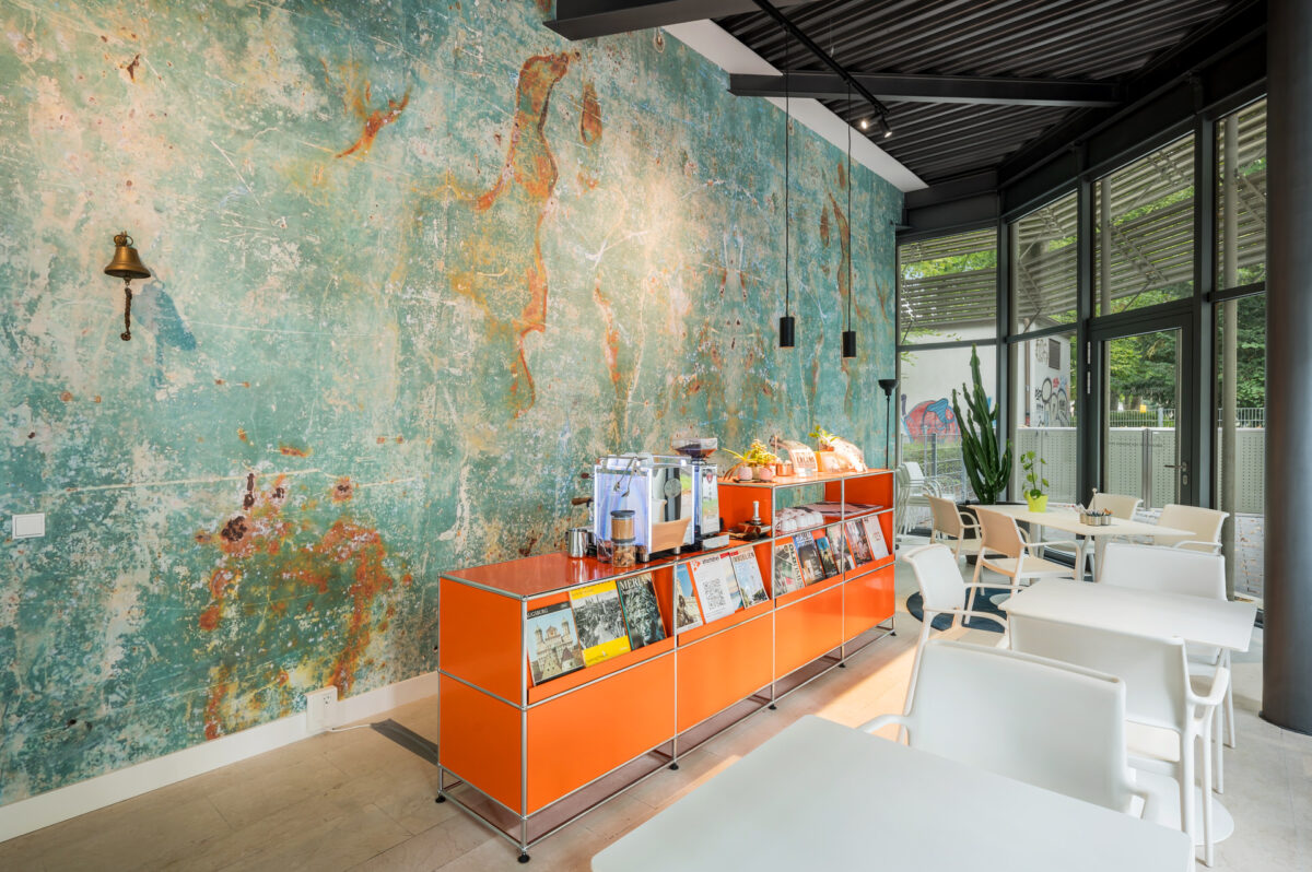 Christian Lorenz Fotograf Ein modernes Café mit weißen Tischen und Stühlen, einer orangefarbenen Theke mit Zeitschriften und Snacks und einer strukturierten Wand in Teal und Orange, neben großen Fenstern mit Blick auf das Grün draußen. Christian Lorenz Fotograf Ein modernes Café mit weißen Tischen und Stühlen, einer orangefarbenen Theke mit Zeitschriften und Snacks und einer strukturierten Wand in Teal und Orange, neben großen Fenstern mit Blick auf das Grün draußen.