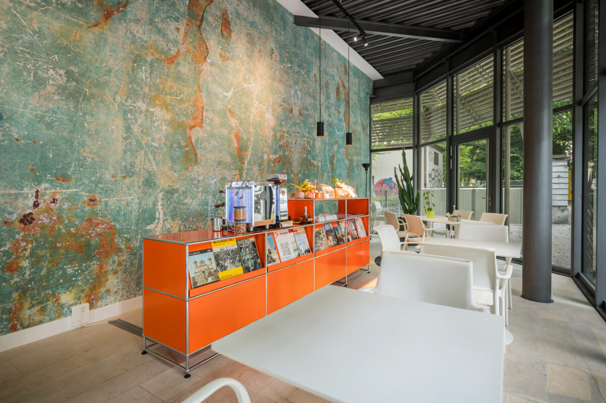 Christian Lorenz Fotograf Ein modernes Café mit weißen Tischen und Stühlen, einer orangefarbenen Vitrine mit Zeitschriften, einer Kaffeemaschine und einer grün strukturierten Wand. Große Fenster lassen natürliches Licht herein und geben den Blick auf das Grün draußen frei. Christian Lorenz Fotograf Ein modernes Café mit weißen Tischen und Stühlen, einer orangefarbenen Vitrine mit Zeitschriften, einer Kaffeemaschine und einer grün strukturierten Wand. Große Fenster lassen natürliches Licht herein und geben den Blick auf das Grün draußen frei.