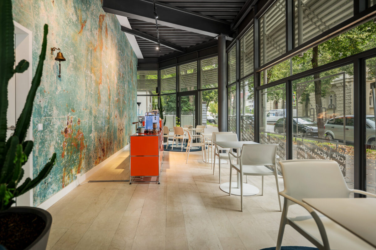 Christian Lorenz Fotograf Modernes Café mit großen, bodentiefen Fenstern, weißen Stühlen und Tischen, einem orangefarbenen Servierwagen, einem grünen Topfkaktus und einer bunt strukturierten Wand. Durch das Glas kann man die Bäume und die Straße sehen. Christian Lorenz Fotograf Modernes Café mit großen, bodentiefen Fenstern, weißen Stühlen und Tischen, einem orangefarbenen Servierwagen, einem grünen Topfkaktus und einer bunt strukturierten Wand. Durch das Glas kann man die Bäume und die Straße sehen.